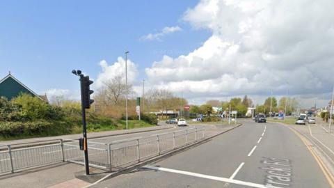 Crossing point on road with traffic lights and metal barriers looking towards a roundabout with vehicles travelling towards on the right and away on the left.