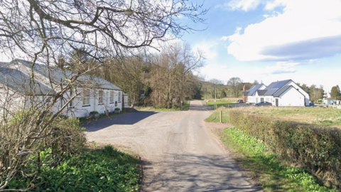 The village, on the left of the picture, is a white-walled single-storey building on a country road. There is a modern, white-walled house further down the single track road. It is a bright, sunny day.
