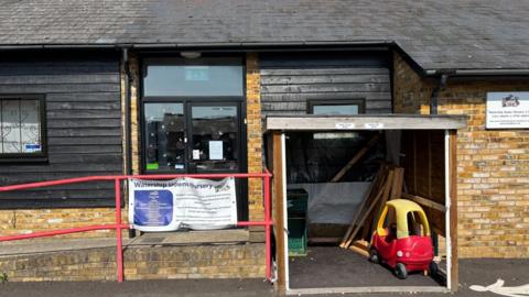 A building with tan bricks and black cladding has signage on the outside saying Watership Downs nursery. A child's red and yellow Cozy Coupe car is in a shed in front of the building.