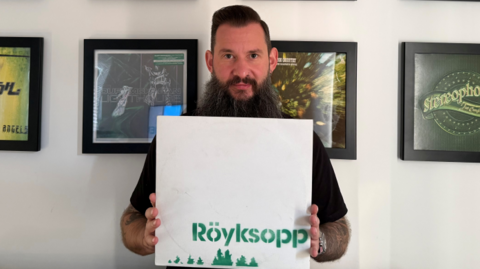 A man with dark hair and a long beard is holding a white vinyl with the word 'Röyksopp' in a green, stencilled format and green pixelated treetops. Behind him is various framed vinyls.