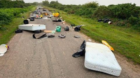 The image shows a long stretch of narrow rural road heavily covered in fly-tipped waste, with rubbish scattered across both the tarmac and the grassy verges on either side. The road is bordered by thick green hedgerows and low bushes, suggesting a quiet countryside location.