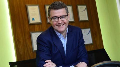 Brendan Mooney smiles while sitting at a desk with a Kainos sign above his head. He has short dark hair and black-framed glasses and is wearing a navy suit jacket and open-necked shirt 