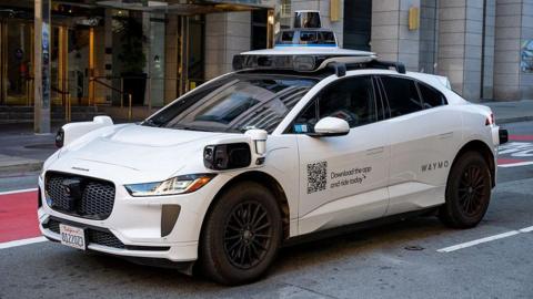 A white Waymo self-driving car with the words Waymo on the side, parked outside a building.