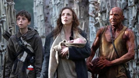 Alfie Williams, Jodie Comer & Ralph Fiennes in a film still from 28 Years Later. They are all looking up and in to the distance at the same thing off camera with worried facial expressions. Jodie Comer is in the centre of the trio and is carrying a baby 