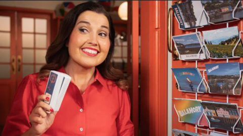 Penny Pocket is a woman in her 40s or 50s with dark hair which curls to her shoulders. She is wearing a red shirt. She is standing at the door to a shop with some postcards hanging from the wall outside.