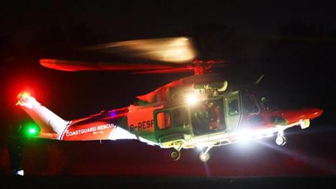 Coastguard helicopter at night with search light on and coastguard team at the door of the aircraft.