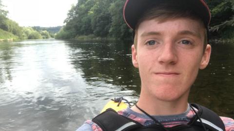 Thomas Garland, who has short ginger hair and freckles and is wearing a red baseball cap, a red and grey t shirt and a life jacket. He is in his canoe, which is not visible, but you can see the water and greenery of the River Wye behind him.