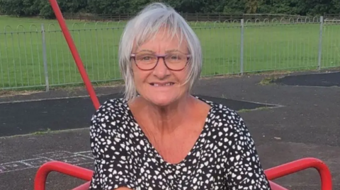 Elizabeth Kennedy sitting on a piece of red playground equipment. She is looking directly at the camera. She has grey hair, which is partially obscuring her face. She is wearing a black top with a small white heart pattern covering it.