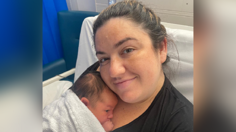 Close up image of Claire smiling whilst holding baby Osian. She is sat in a hospital bed. Clare has dark brown hair and is wearing a black t-shirt. Osian is wrapped in a towel and is resting on her chest. 
