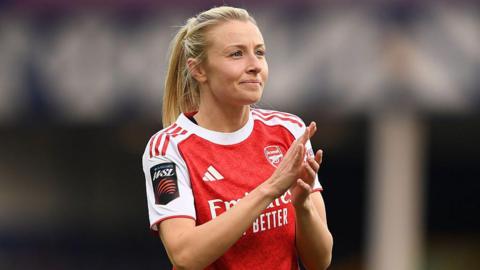 Leah Williamson, in a red Arsenal shirt with white sleeves and blonde hair tied back in a ponytail, looks in the direction of the crowd and applauds