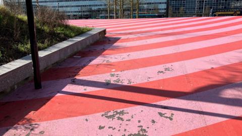 A wide outdoor pathway painted with alternating pink and red diagonal stripes, showing areas of wear. Shadows from nearby poles stretch across the surface, with a low concrete border and some greenery on the left. In the background, leafless trees stand in front of Milton Keynes railway station.