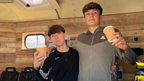Corey (left) and Deacon (right), are standing inside a small kitchen in a converted horse box. They are both holding paper coffee cups. Both have short, brown hair. Corey is wearing a black top and Deacon is wearing a two-tone brown top.