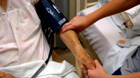 A nurse is taking the blood pressure of an older patient who is wearing a hospital gown and is sat alongside a hospital bed