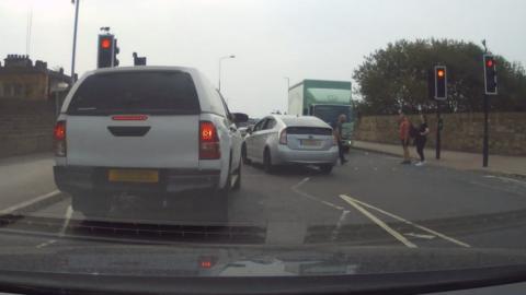 A van and a lorry stopped at traffic lights. A car has driven through a red light, and there are people crossing at the lights.