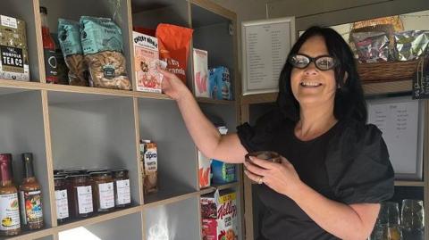 Rachael Parke is standing in her community shop in front of shelves of food. She has shoulder length black hair, wears black glasses and is holding a mug of coffee and a box of cereal. 