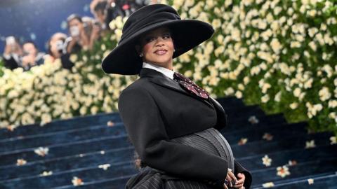 Rihanna appears at the 2025 Met Gala visibly pregnant.  She is wearing a black and grey tailored outfit and large brim hat. 