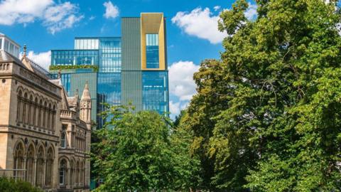 A photograph of the skyline where the new design and digital arts building at Nottingham Trent University can be seen in the distance. It's a tall glass building with a section of windows at the top surrounded by a yellow border. 