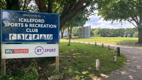 A blue and white sign for Ickleford Sports & Recreation Club advertising Sky Sports and BT Sport. A path can also be seen leading to football and cricket pitches.