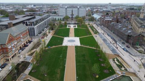 An aerial view of Queen’s Gardens in Hull shows long, symmetrical lawns divided by wide pathways that converge on a central circular plaza, framed by newly planted trees. The gardens are surrounded by a mix of modern and historic buildings and the city stretching out into the distance beyond.