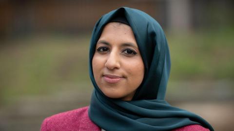 Zara Mohammed looks into the camera with a small smile. She wears a dark pink jacket and a teal green hijab.