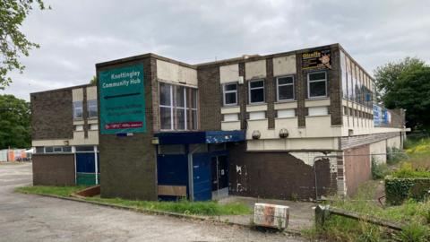 A visibly decrepit brutalist building with sharred paintwork. The grass around it looks overgrown.
