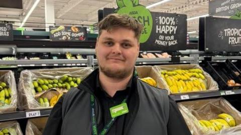 Phil Snow-Griffiths has brown hair and a beard and is wearing an Asda lanyard and uniform. He stands in front of the fruit and veg section.