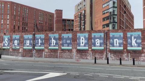 The words 'South Bank' are written in large white writing on top of blue boards stuck to a red brick wall. Multi-storey buildings are behind the wall, and there is an empty road in front of it.