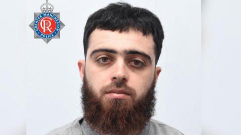 A mugshot of Mohammed Jaza Hamasaleh including the Greater Manchester Police logo. Hamasaleh has short dark hair and a long, thick beard and is wearing grey clothes. 