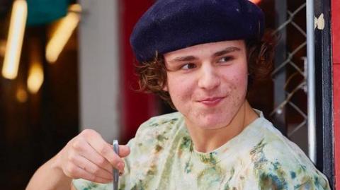 A teenage boy wearing a navy beret and a green tie-dye T-shirt. He has curly brown hair that reaches the bottom of his ears
