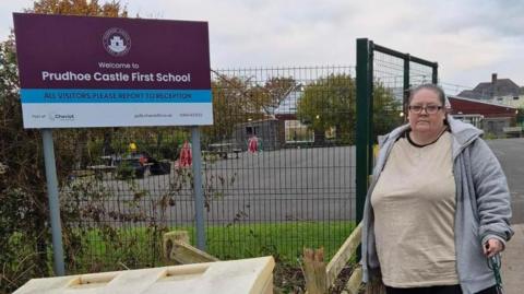Michelle Gallon is wearing a grey hoodie and a white top. She has glasses and has hair tied back. She is holding a walking stick. She is standing by a large purple sign which says "Prudhoe Castle First School". It is daytime. School grounds can be seen in the background with tables. 