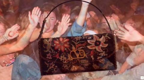 A screenshot from Valentino's AI-generated video shared on Instagram of a black, embroidered edition of its DeVain bag. It is dangling in front of a crowd of people whose bodies and faces blurring into each other.