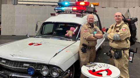 Two shaven-headed men are standing next to a white car which has blue and red lights on its roof. The men are wearing light brown all-in-one Ghostbuster suits and have their arms crossed. In front of them is a round white table which has the print of a white ghost that is climbing out of a red and black 'no entry' sign - the logo of the Ghostbusters film.