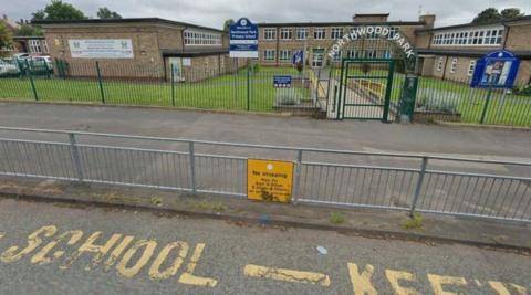 Yellow road markings are on the road in the foreground. The school is behind a fence and the words Northwood Park are above the gate that forms the entrance to the grounds.