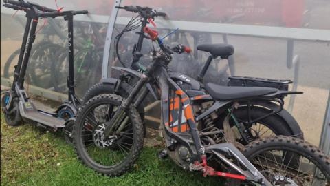 Several e-scooters, e-bikes and off-road bikes. Most are coloured black, and one of the bikes has an orange pattern and orange handles. They are stood on grass leaning against a clear perspex bike shelter, through which other bikes can be seen in the background.