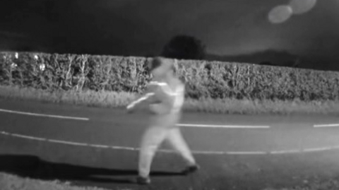 A blurred image of a man walking past a house at night with the road in the background. The image was taken from Ring doorbell footage.