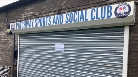 The closed metal shutters of Patchway Sports and Social Club. There is a paper sign on the shutters saying 'closed as of immediate effect' 