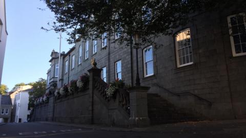 Guernsey government building. It is a grey stone building which has a stone staircase leading up to it, which has flowers draped over it. There is a road running in front of the building.