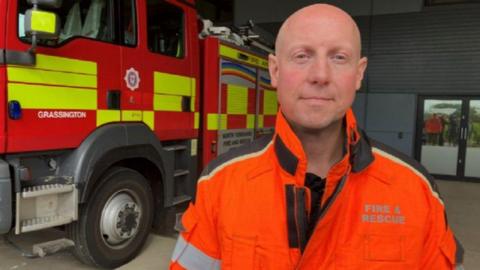Jonathan Dyson stood in front of a fire engine wearing an orange and black jacket that says "fire and rescue" on it. He has a bald head and is looking at the camera with a slight smile.