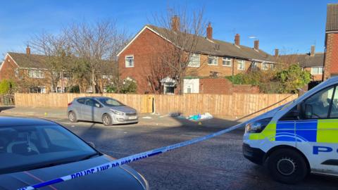 The scene of the stabbing. Police tape has been put up across the road. A police van is parked in the middle of the road to the right of the picture. A pile of items, which look like rubbish, are on the pavement by a garden fence. Yellow police markers have been placed on the pavement near them.