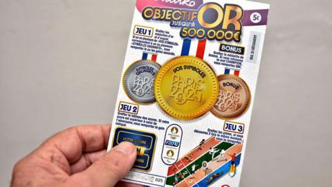 A man's hand holding a French scratch card which is Paris Olympics themed. 