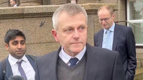 Deputy Philip Ozouf is pictured outside the Royal Court. Two men are following him. All the people in the photo are wearing suits.