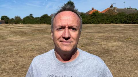 A man with his arms behind his back in a grey T-shirt, he has light brown short hair and behind him is an undulating area of very dry and almost dead grass caused by heat and no rain. A few houses can be seen in the far distance.