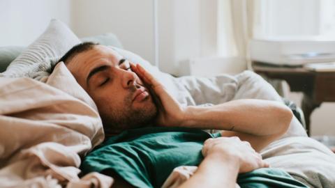 A man lays in amongst cushions and blankets, his eyes are closed and he looked fatigued.