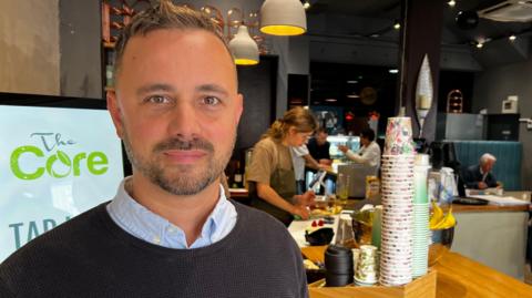 Kris Talikowski stands in a cafe with disposable cups piled up behind him. Staff are preparing food and a white-haired man is eating in a booth in the background.