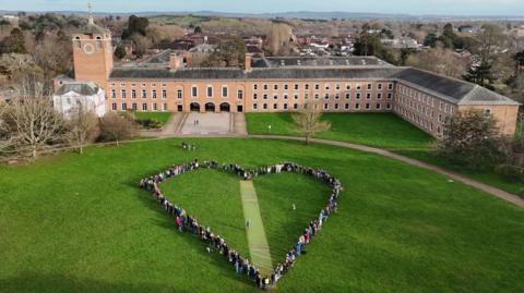 A crowd of people forming a heart shape around the cricket pitch on the grounds of County Hall with County Hall, a large L-shaped building with a tower to the left of the image, in the background.