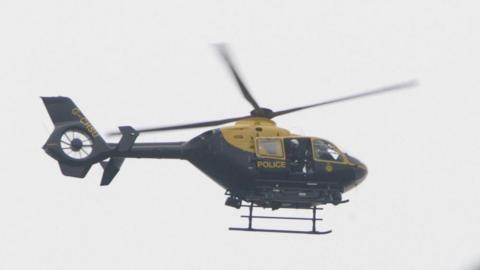 A photograph of a yellow and black police helicopter in the air. There is a person slightly leaning through the door wearing a white helmet.