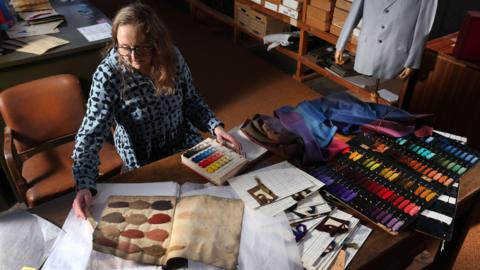 Image taken from above of a woman sat at a large desk, she is wearing a light blue and black patterned shirt and has medium length brown hair and glasses. In front of her are dye ledgers and papers as well as a range of coloured dye samples.