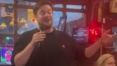 Man in black sweater closes his eyes as he sings into microphone - a woman in a white shirt with blonde hair sits behind him. You can see the words on karaoke machine.