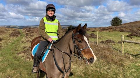 A woman wearing a hi-viz tabard and a riding hat, sitting on a brown, clipped horse, with the Shropshire hills behind.