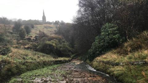 A reservoir spillway leading down hill, with bushes and grass to each side. A church tower can be seen on the distant cloudy skyline.